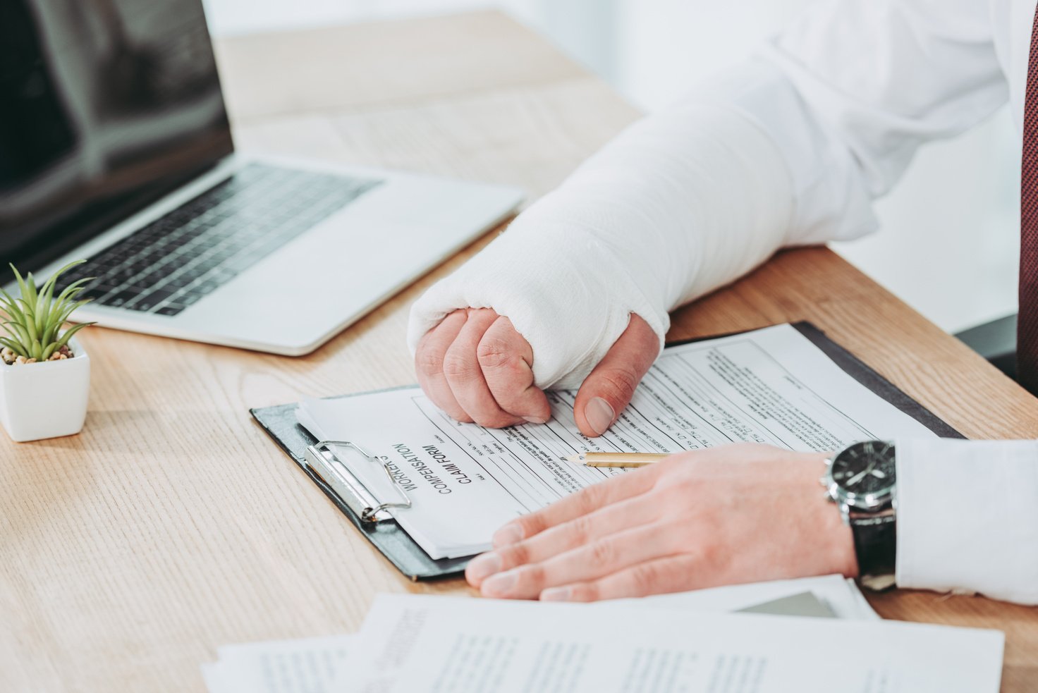 cropped view of worker with broken arm in gypsum sitting at table and holding hands on compensation form in office, compensation concept